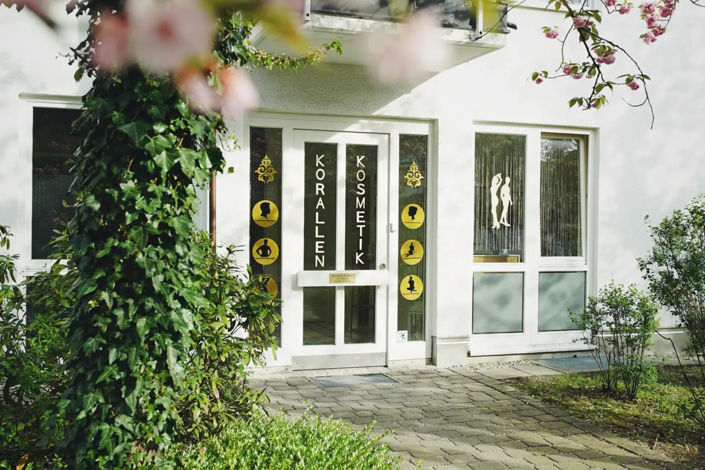 Eingang von Kosmetik Korallen in der Kornwegerstraße 59 in München-Hadern, umrahmt von Efeu und einem blühenden Baum