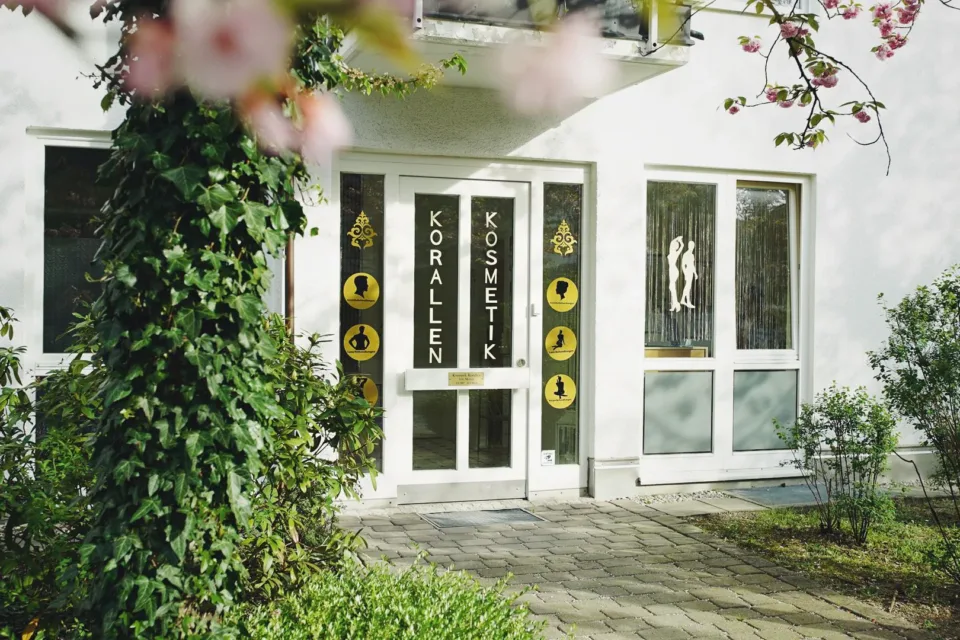 Eingang von Kosmetik Korallen in der Kornwegerstraße 59 in München-Hadern, umrahmt von Efeu und einem blühenden Baum