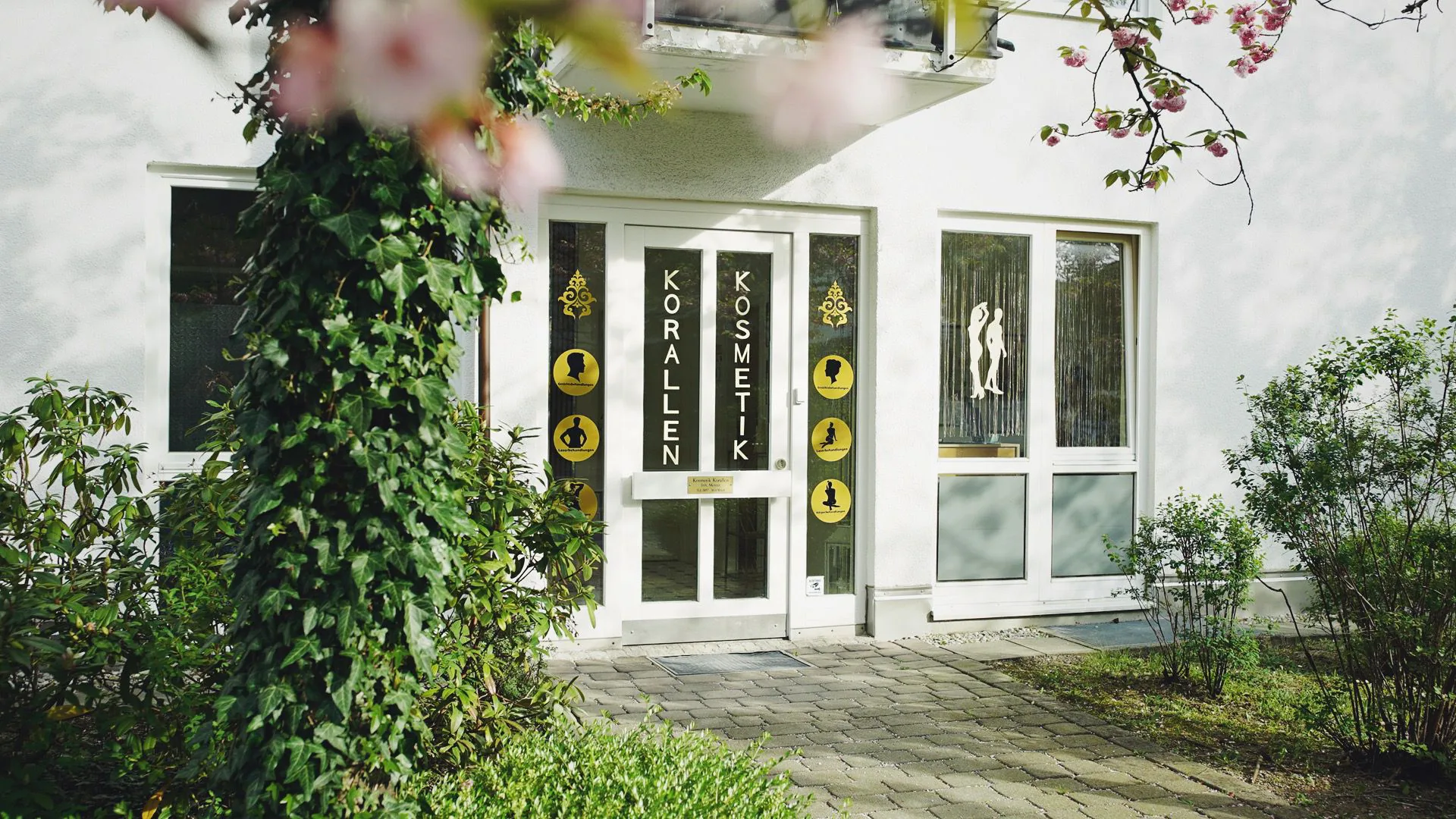 Eingang von Kosmetik Korallen in der Kornwegerstraße 59 in München-Hadern, umrahmt von Efeu und einem blühenden Baum