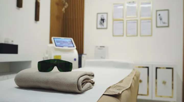 Laserschutzbrille auf einer gefalteten Decke auf der Behandlungsliege, dahinter das Lasergerät für die Haarentfernung